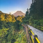 Pathway with yellow-and-black railing winds along a lush forested canyon, a volcano rises in the distance under a blue sky.