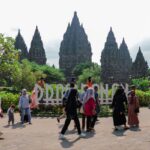 Tourists walk past a large Prambanan sign with towering ancient temple spires in the background and greenery around.