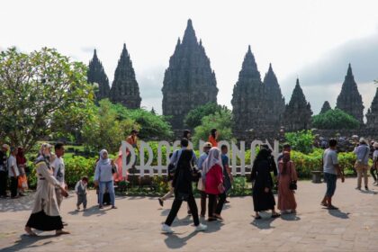 Tourists walk past a large Prambanan sign with towering ancient temple spires in the background and greenery around.