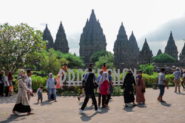 Tourists walk past a large Prambanan sign with towering ancient temple spires in the background and greenery around.