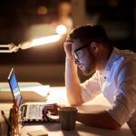 Man in a white shirt works at a laptop late at night, hand on head, glasses reflecting the screen in a dim office.