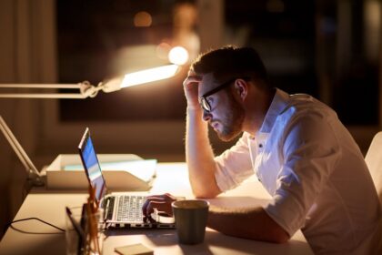 Man in a white shirt works at a laptop late at night, hand on head, glasses reflecting the screen in a dim office.
