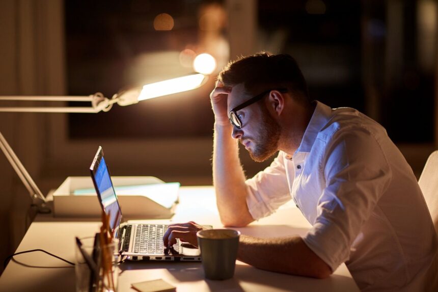 Man in a white shirt works at a laptop late at night, hand on head, glasses reflecting the screen in a dim office.