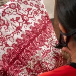 Close-up of a person from behind, wearing red, handling a red-and-white batik fabric with intricate floral patterns.
