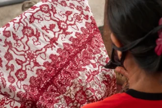 Close-up of a person from behind, wearing red, handling a red-and-white batik fabric with intricate floral patterns.