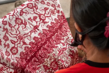 Close-up of a person from behind, wearing red, handling a red-and-white batik fabric with intricate floral patterns.