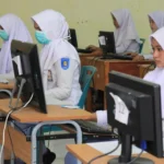 Group of girls in white uniforms and hijabs using computers in a classroom; some wear surgical masks and badges on sleeves.