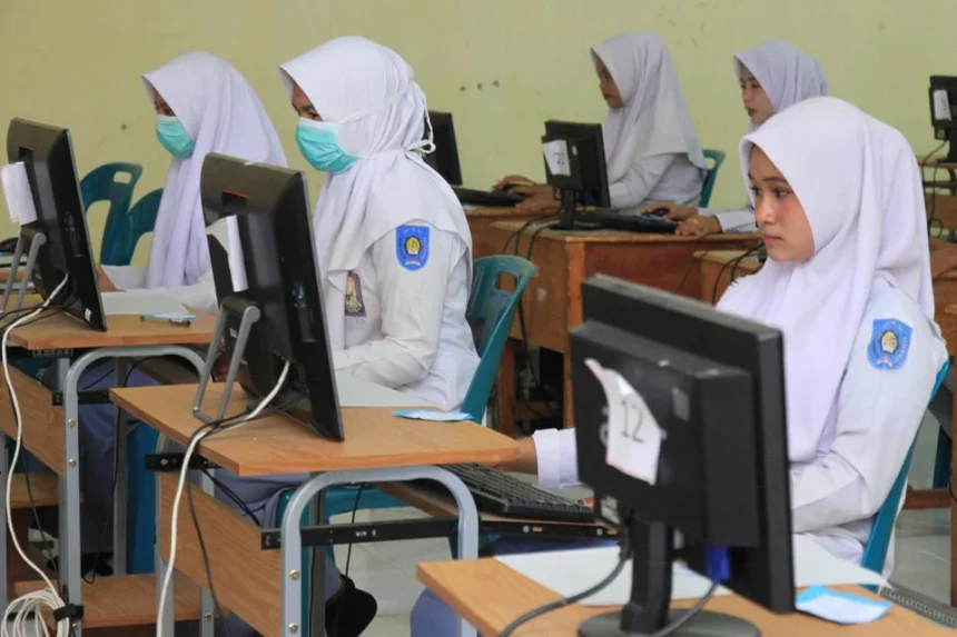 Group of girls in white uniforms and hijabs using computers in a classroom; some wear surgical masks and badges on sleeves.