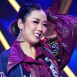 Smiling woman performing on stage with maroon patterned jacket and large geometric earrings, under blue and yellow stage lights.