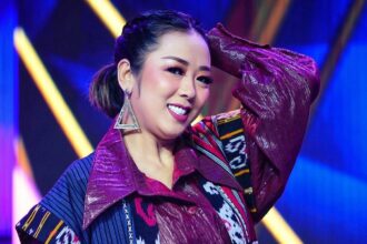 Smiling woman performing on stage with maroon patterned jacket and large geometric earrings, under blue and yellow stage lights.