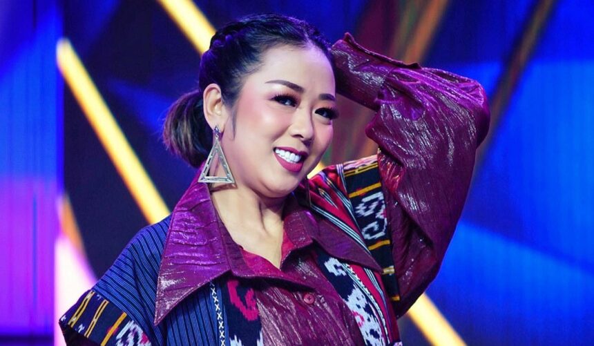 Smiling woman performing on stage with maroon patterned jacket and large geometric earrings, under blue and yellow stage lights.