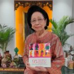 Elderly woman in a peach blouse and glasses holding a colorful book titled '95 Perempuan Tangguh Inspiratif' indoors with plants in the background.