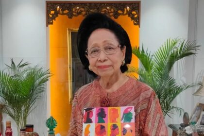 Elderly woman in a peach blouse and glasses holding a colorful book titled '95 Perempuan Tangguh Inspiratif' indoors with plants in the background.