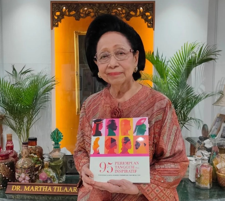 Elderly woman in a peach blouse and glasses holding a colorful book titled '95 Perempuan Tangguh Inspiratif' indoors with plants in the background.