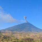 Active volcano spews a small ash plume from its crater under a clear blue sky, with dry scrubland in the foreground.