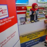 Teller assists a customer at a service counter in a red-and-white branded co-op, with 'Koperasi Kelurahan Merah Putih' signage in the foreground.