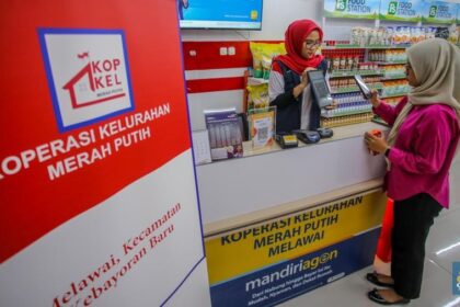 Teller assists a customer at a service counter in a red-and-white branded co-op, with 'Koperasi Kelurahan Merah Putih' signage in the foreground.