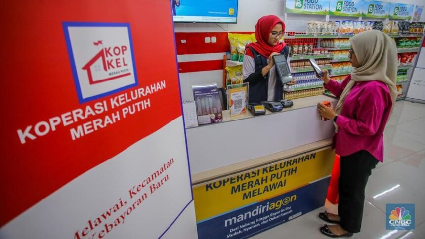 Teller assists a customer at a service counter in a red-and-white branded co-op, with 'Koperasi Kelurahan Merah Putih' signage in the foreground.