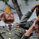 Veterans in olive uniforms salute during a ceremony, wearing yellow/gold caps.