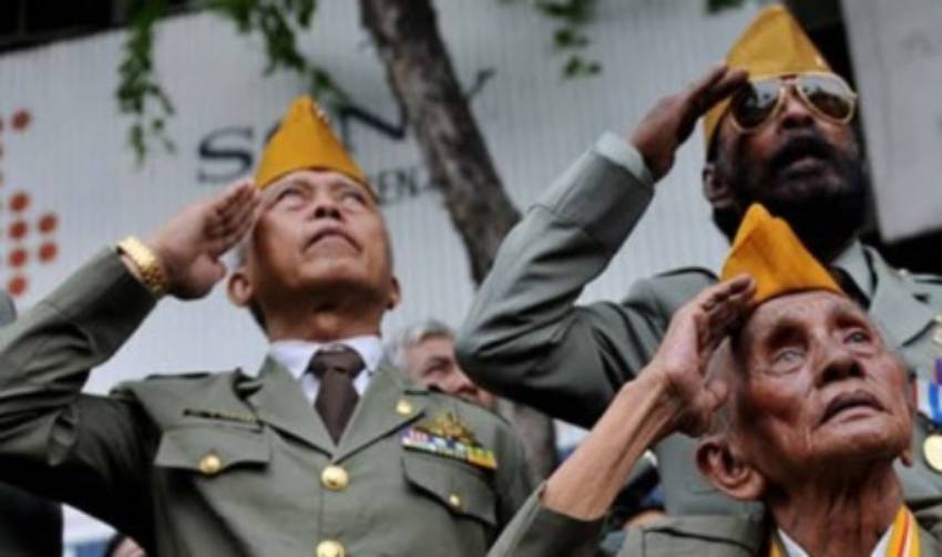 Veterans in olive uniforms salute during a ceremony, wearing yellow/gold caps.