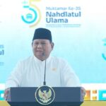 Speaker in a white outfit and black peci delivers a speech at a podium with the Nahdlatul Ulama seal during the 35th Muktamar event, blue backdrop in view.