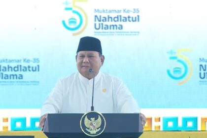 Speaker in a white outfit and black peci delivers a speech at a podium with the Nahdlatul Ulama seal during the 35th Muktamar event, blue backdrop in view.