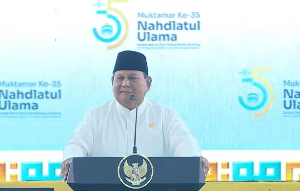 Speaker in a white outfit and black peci delivers a speech at a podium with the Nahdlatul Ulama seal during the 35th Muktamar event, blue backdrop in view.