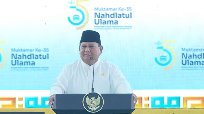 Speaker in a white outfit and black peci delivers a speech at a podium with the Nahdlatul Ulama seal during the 35th Muktamar event, blue backdrop in view.