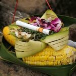 Banana leaf plate holding corn on the cob, folded leaves, purple flowers, and a cigarette resting on top.