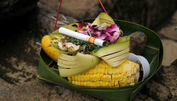Banana leaf plate holding corn on the cob, folded leaves, purple flowers, and a cigarette resting on top.