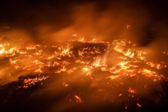 Nighttime wildfire burning through dry vegetation with bright flames and rising smoke across a scorched landscape