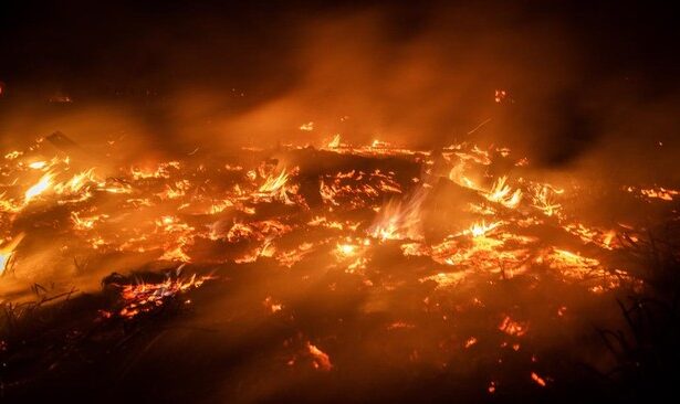 Nighttime wildfire burning through dry vegetation with bright flames and rising smoke across a scorched landscape