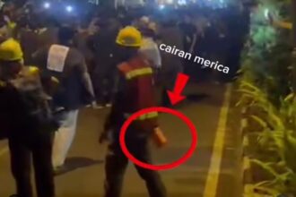Nighttime street scene with workers in yellow helmets and a crowd, a person in the foreground is circled, holding a small orange object in their hand while others watch.
