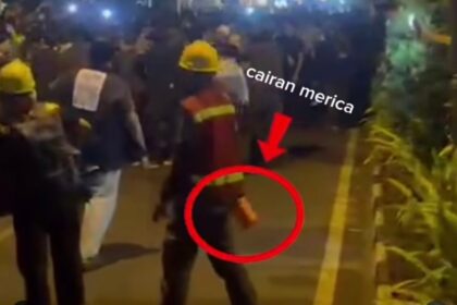 Nighttime street scene with workers in yellow helmets and a crowd, a person in the foreground is circled, holding a small orange object in their hand while others watch.