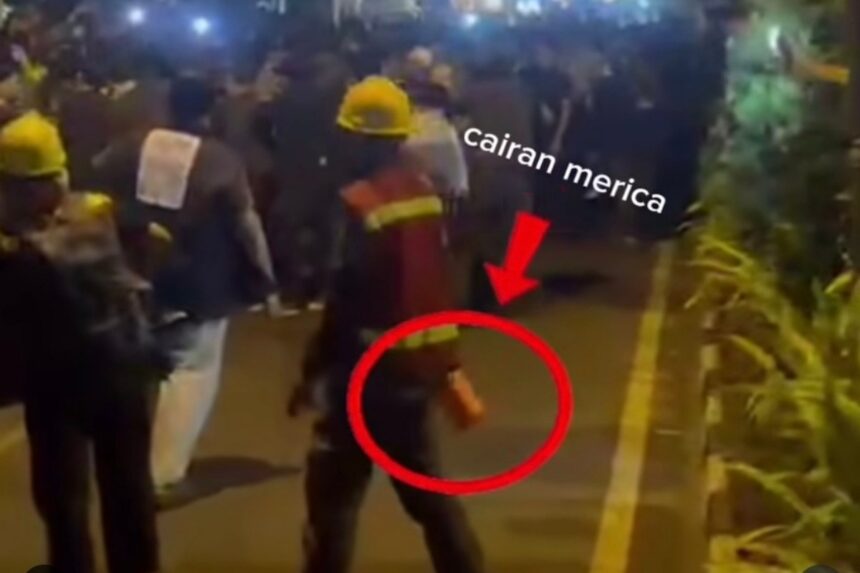 Nighttime street scene with workers in yellow helmets and a crowd, a person in the foreground is circled, holding a small orange object in their hand while others watch.