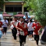 Group of students in red-and-white uniforms run toward the camera in a schoolyard, with a staff member in a high-visibility vest and security personnel visible in the foreground.