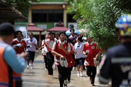Group of students in red-and-white uniforms run toward the camera in a schoolyard, with a staff member in a high-visibility vest and security personnel visible in the foreground.