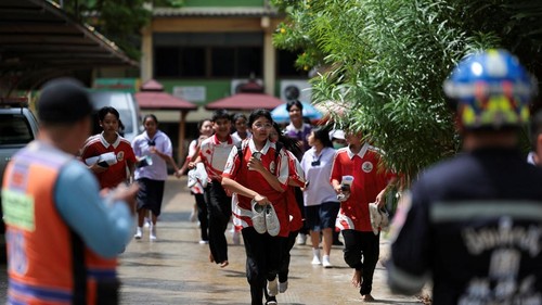 Group of students in red-and-white uniforms run toward the camera in a schoolyard, with a staff member in a high-visibility vest and security personnel visible in the foreground.