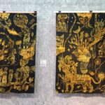 Two black-and-gold tapestries with intricate yellow drawings hanging on a gallery wall, each with a small placard beside it.