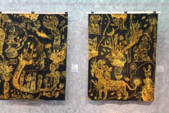 Two black-and-gold tapestries with intricate yellow drawings hanging on a gallery wall, each with a small placard beside it.