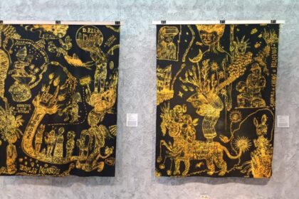Two black-and-gold tapestries with intricate yellow drawings hanging on a gallery wall, each with a small placard beside it.