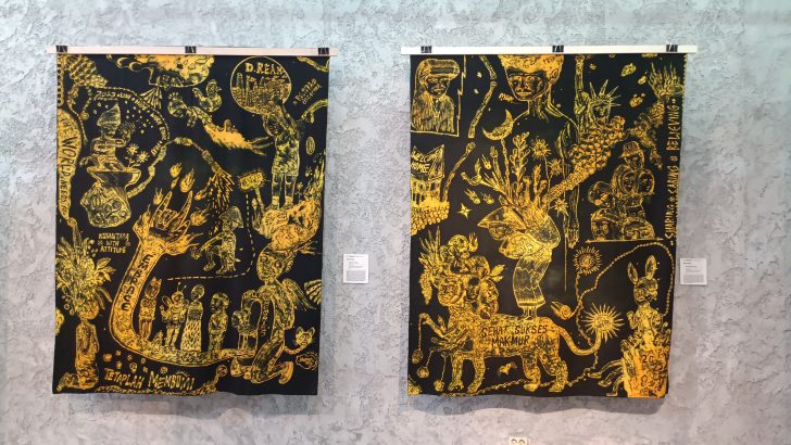 Two black-and-gold tapestries with intricate yellow drawings hanging on a gallery wall, each with a small placard beside it.