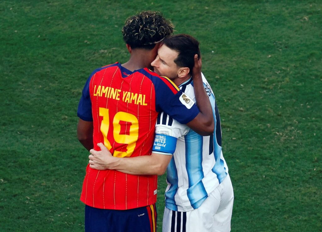 yamal hugging messi by Getty Images - Mabur.co Two soccer players in a hug on a grassy field, one in a red jersey with yellow numbers, the other in a blue-and-white striped kit.