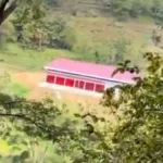 Red-roofed, single-story building with a row of openings in a green hillside, viewed through trees.