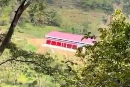 Red-roofed, single-story building with a row of openings in a green hillside, viewed through trees.