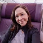 Woman taking a selfie in an airplane cabin, sitting in a purple seat and smiling at the camera with headrest and window area nearby