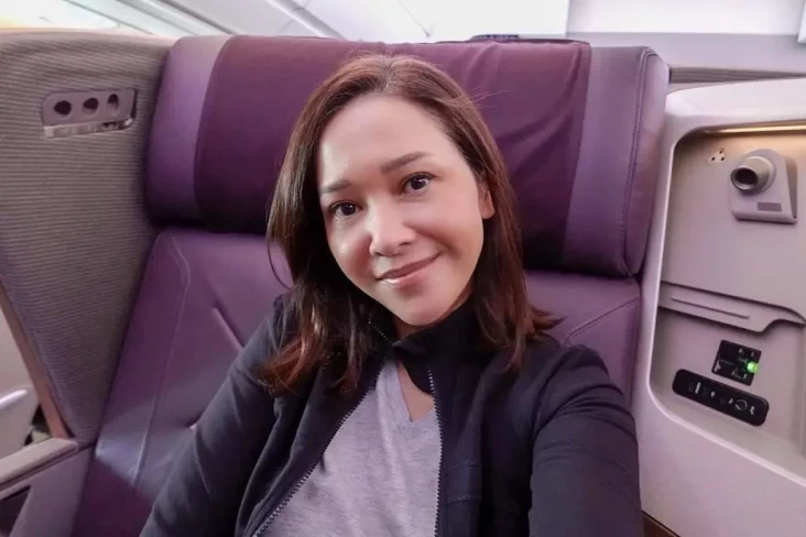 Woman taking a selfie in an airplane cabin, sitting in a purple seat and smiling at the camera with headrest and window area nearby