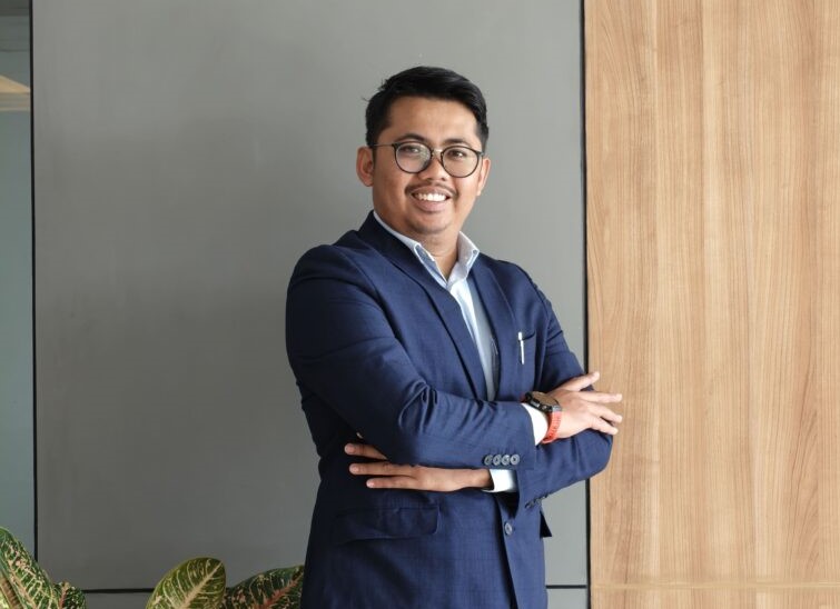 Portrait of a man in a navy suit with glasses, smiling, arms crossed indoors beside a wooden panel.