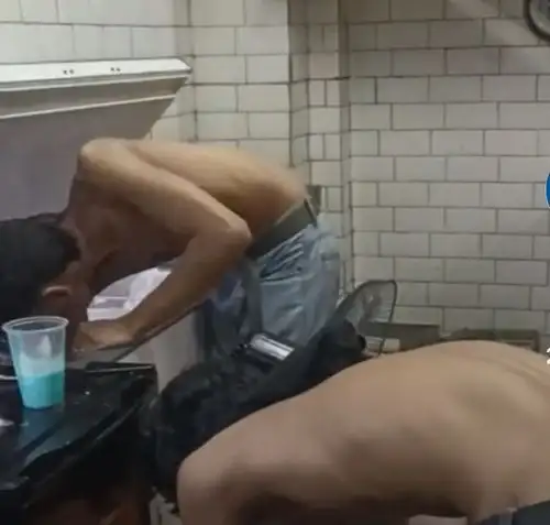 Two shirtless men bent over a counter in a tiled room, with a blue cup nearby.