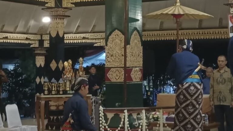 A man in traditional clothing sits near a decorated pillar at a ceremonial event with trophies on a table nearby.